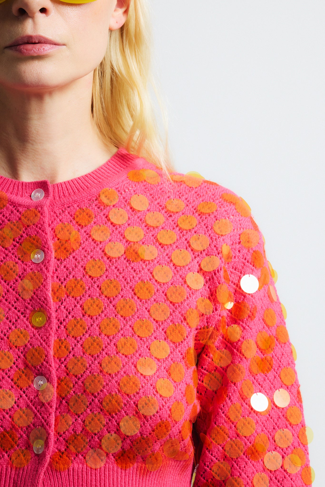 Link to "Mina" cardigan. Image is of a female model wearing cropped bright pink textured cardigan with see-through iridescent orange paillettes. Model is also wearing green crinkle cotton metallic see-through shorts and yellow sunglasses. 