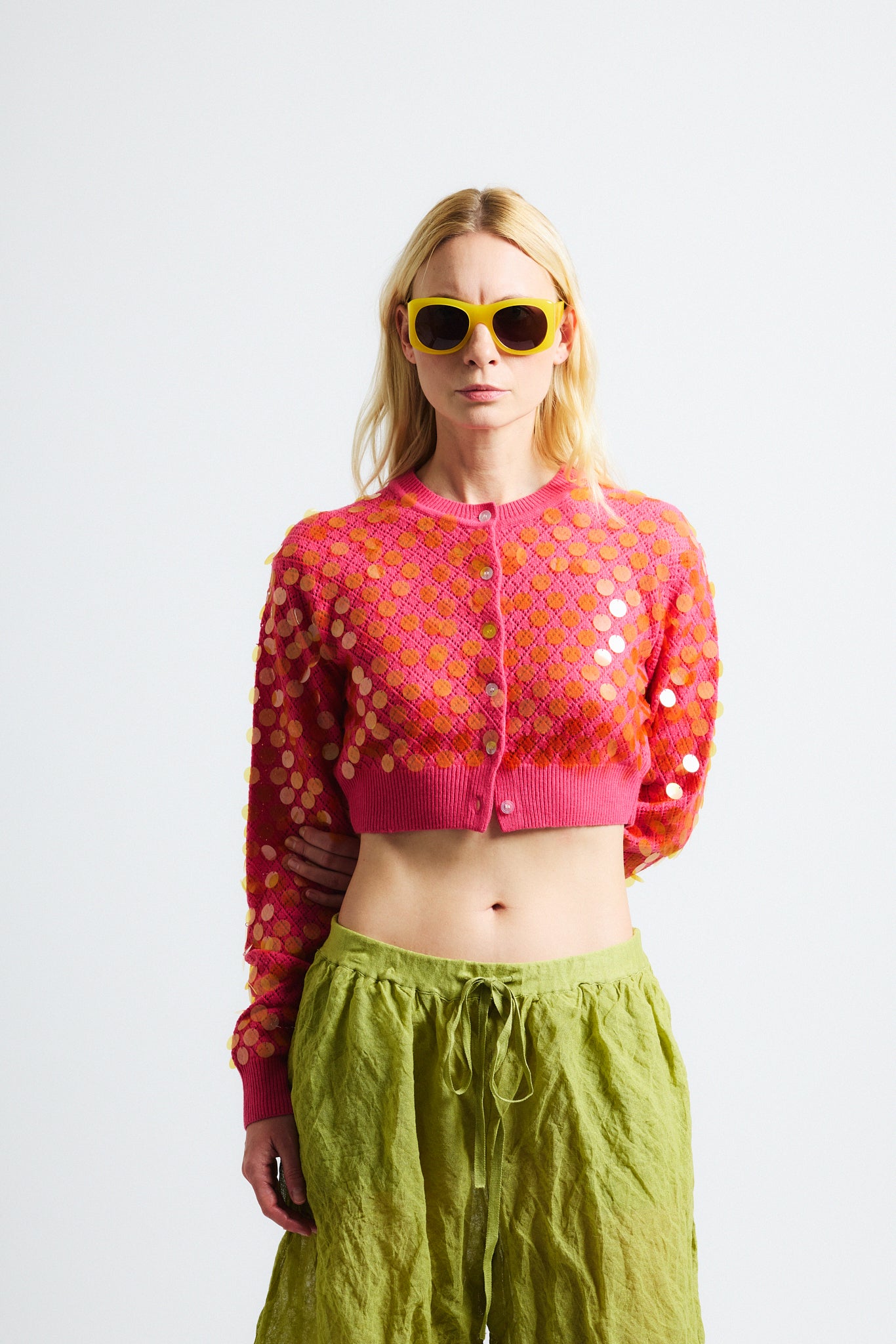 Link to "Mina" cardigan. Image is of a female model wearing cropped bright pink textured cardigan with see-through iridescent orange paillettes. Model is also wearing green crinkle cotton metallic see-through shorts and yellow sunglasses. 