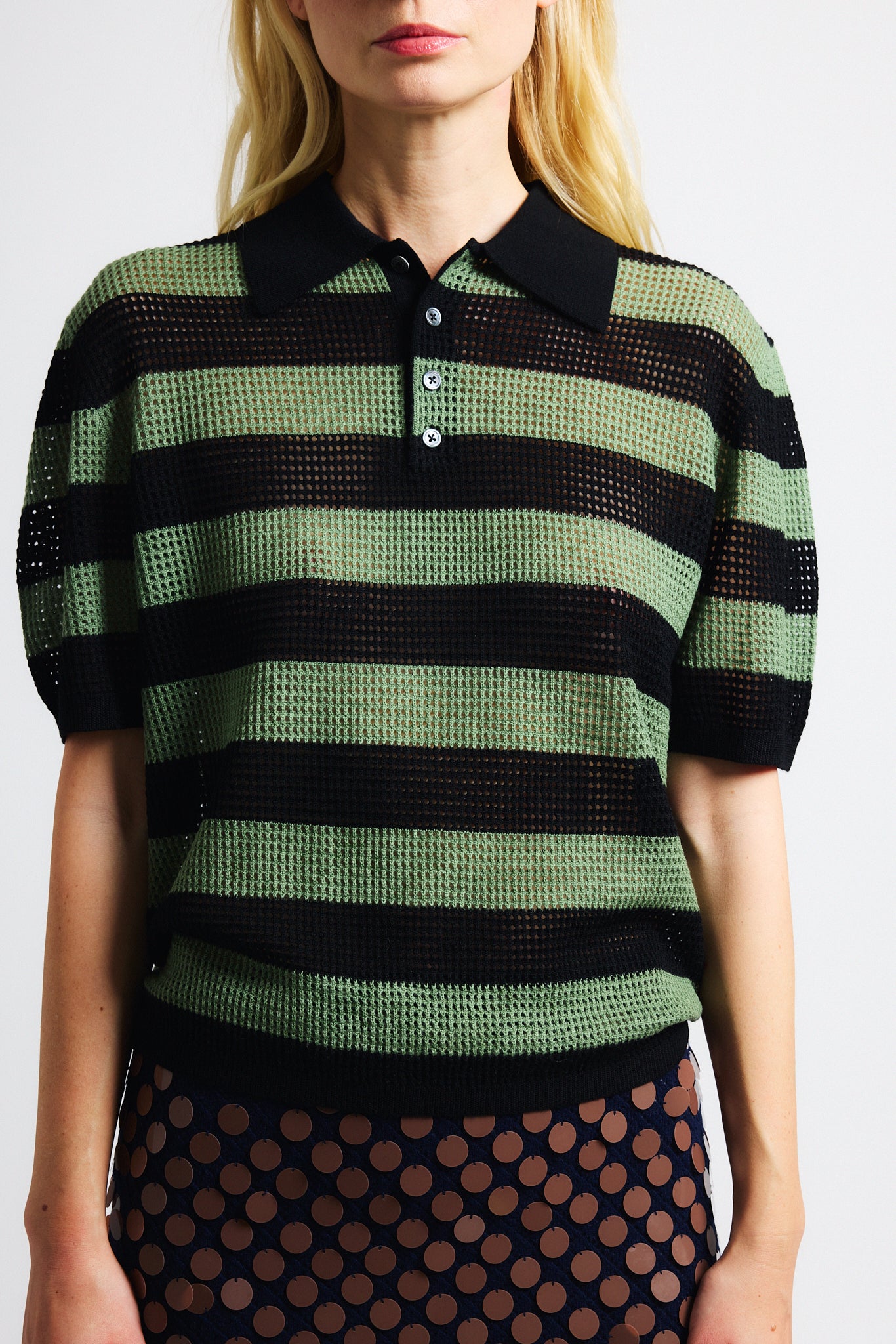 Link to "Luke" polo. Image is of a female model wearing green and black perforated stripe polo shirt and orange sunglasses.