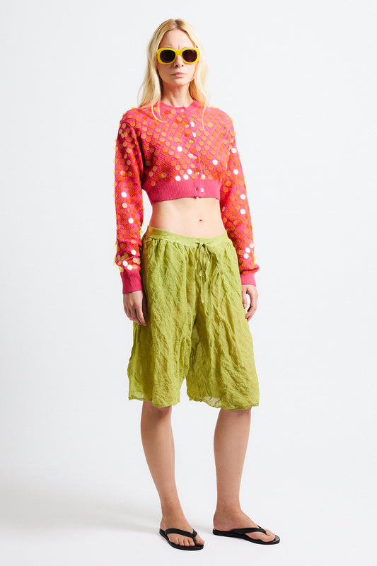 Link to "Mina" cardigan. Image is of a female model wearing cropped bright pink textured cardigan with see-through iridescent orange paillettes. Model is also wearing green crinkle cotton metallic see-through shorts and yellow sunglasses. 