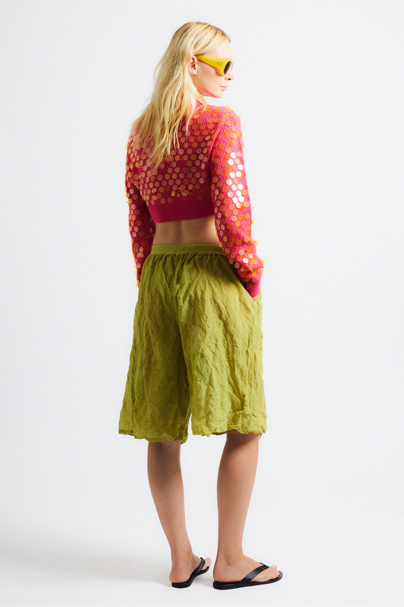 Link to "Mina" cardigan. Image is of a female model wearing cropped bright pink textured cardigan with see-through iridescent orange paillettes. Model is also wearing green crinkle cotton metallic see-through shorts and yellow sunglasses. 