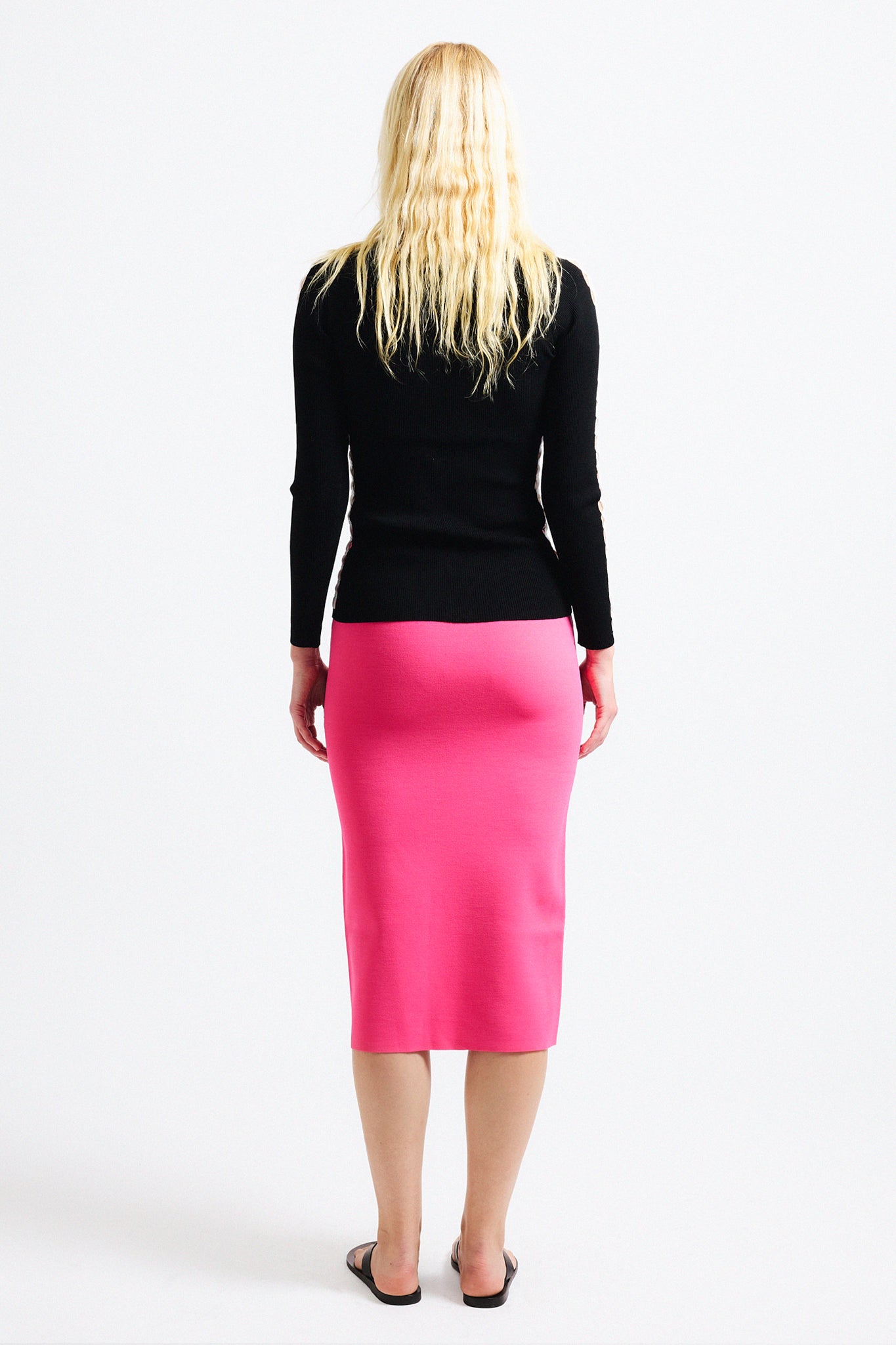 Link to "Issimo" long sleeve. Image is of a female model wearing black think ribbed fitted long sleeve top and pink pencil skirt. The top has a thin stripe of lattice work along the arm/side hems that exposes skin.