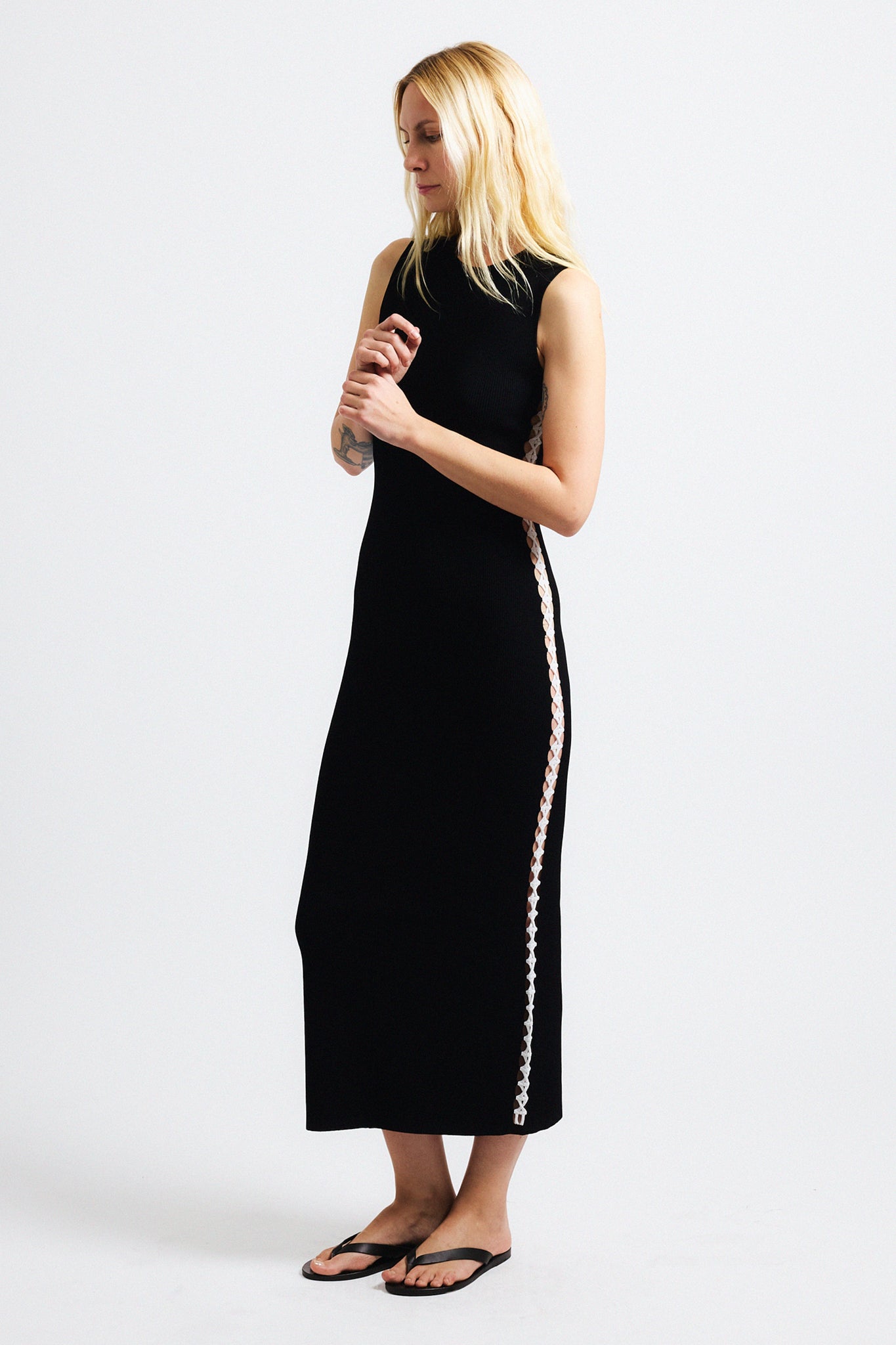 Link to "Hydra" dress. Image is of a female model wearing sleeveless black think ribbed fitted long dress. The dress has a thin stripe of lattice work along the side hems that exposes skin.