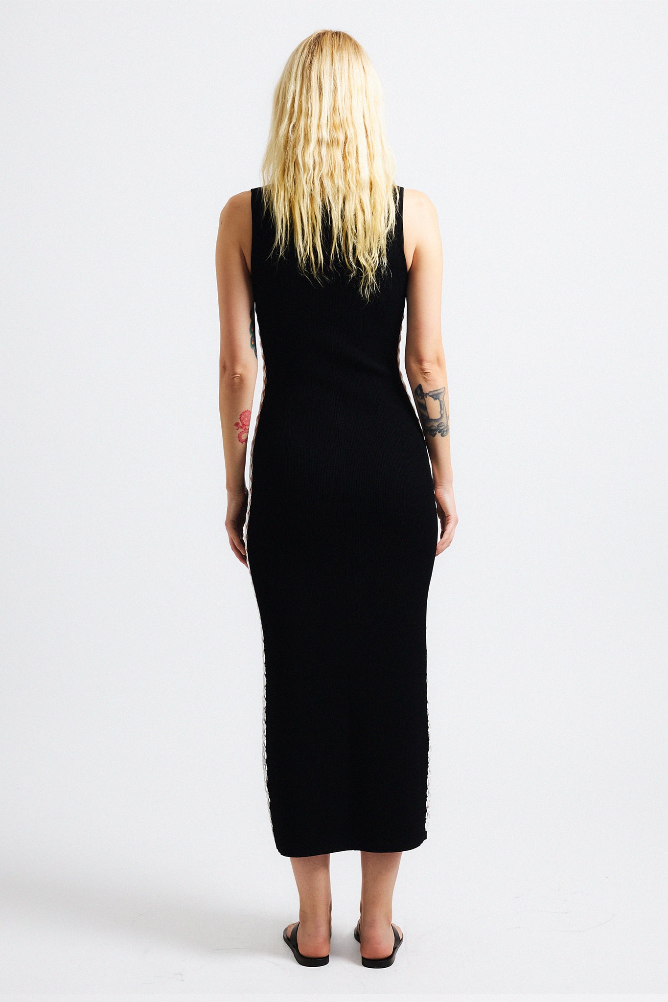 Link to "Hydra" dress. Image is of a female model wearing sleeveless black think ribbed fitted long dress. The dress has a thin stripe of lattice work along the side hems that exposes skin.