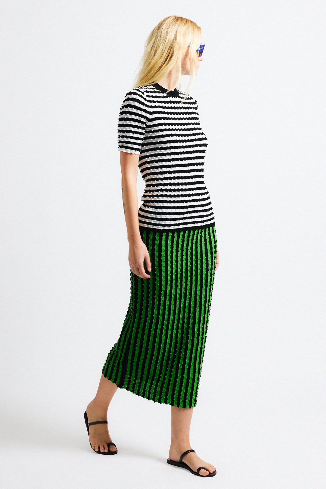 Link to "Capucine" skirt. Image is of a female model wearing black and white horizontal stripe knit crewneck shirt and vertical black/green stripe long skirt. Both pieces have a raised origami texture.